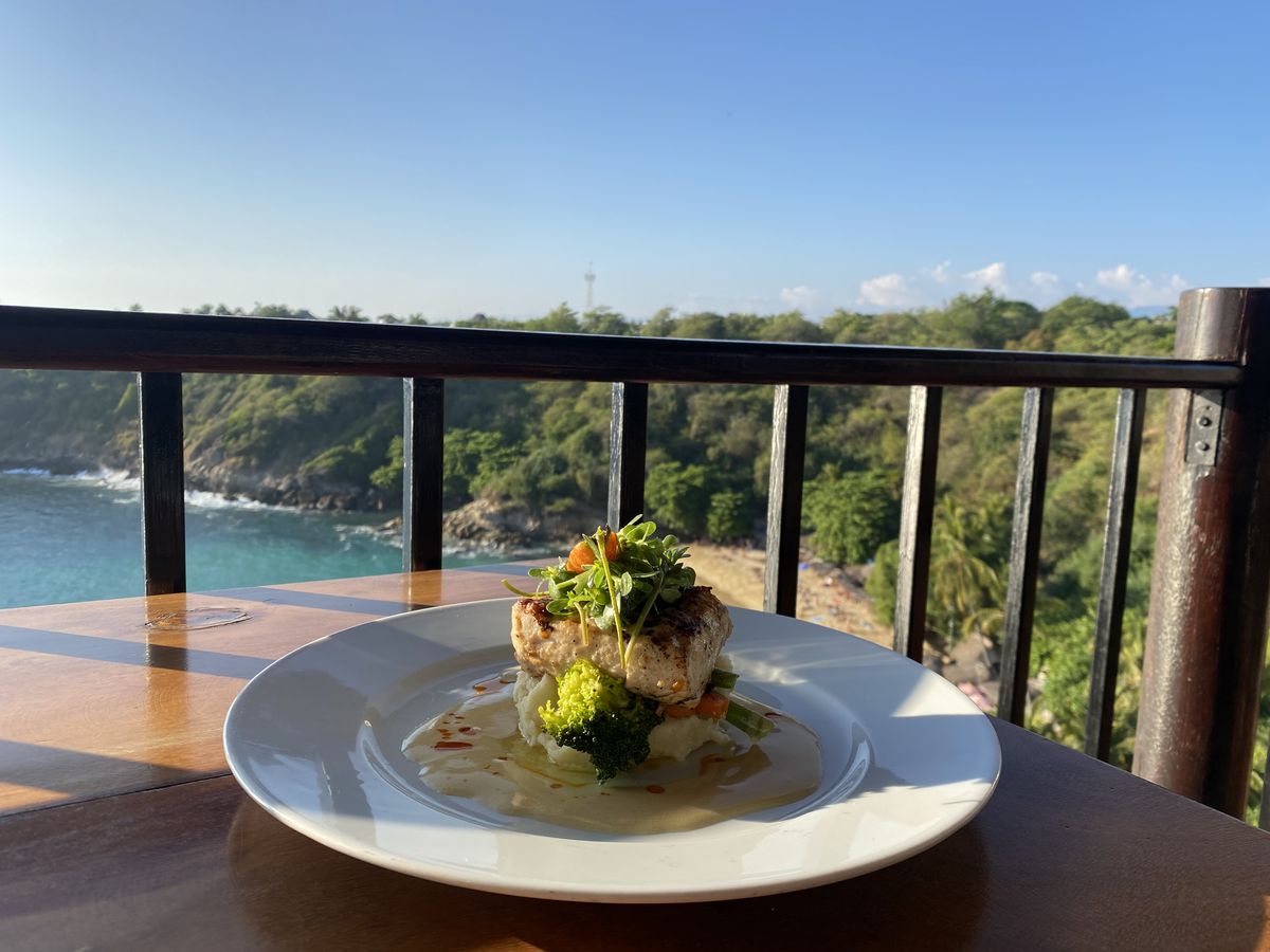 A hunk of fish plated on a mound of mashed potato with vegetables, with a view of a sunny cove in the background