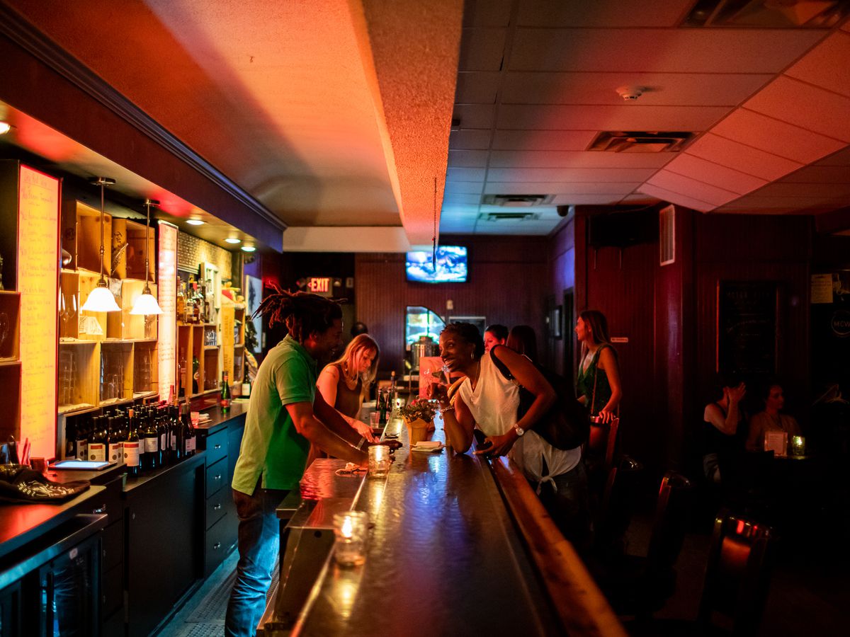a black bartender with a green shirt and dreadlocks tied back leans over a long dark bar to chat with a black woman with short hair and a white shirt. A glowing red menu display casts red light over the drop ceiling.