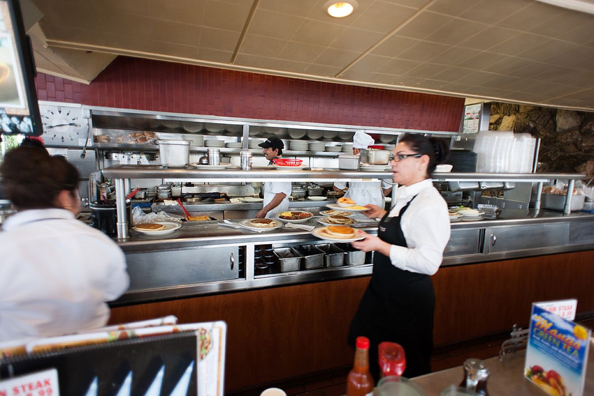 A worker inside of a restaurant carries plates and picks up more from the kitchen in a fast-paced environment.
