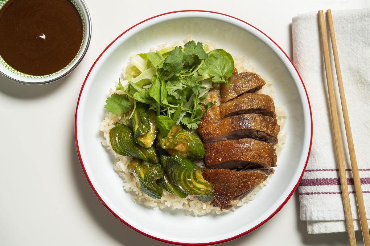 A plate with slices of duck and greens on it with a set of chopsticks laid on a cloth napkin nearby and a cup of dipping sauce at the top left of the photo