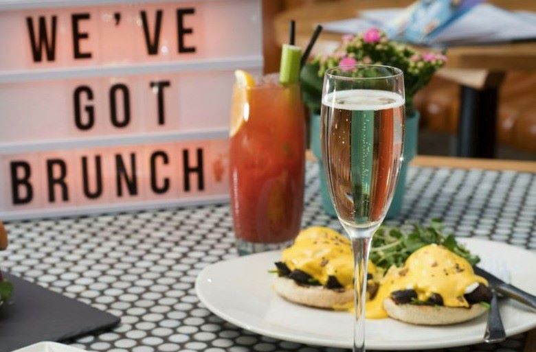 A sign in the background reads “We’ve got brunch.” In the foreground, a flute of sparkling and a white plate topped with fresh baby arugula and poached eggs atop English muffins.