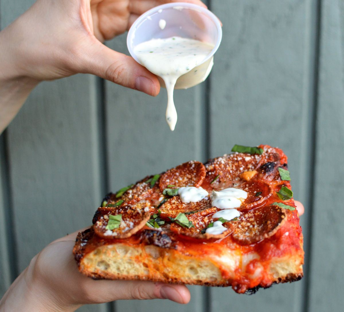 A person pours ranch over a slice of square pizza from Ranch Pizza
