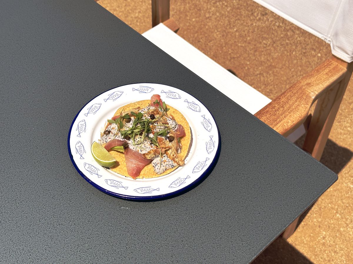 A tostada sits at the edge of a slate-colored table at a daytime Mexican restaurant.
