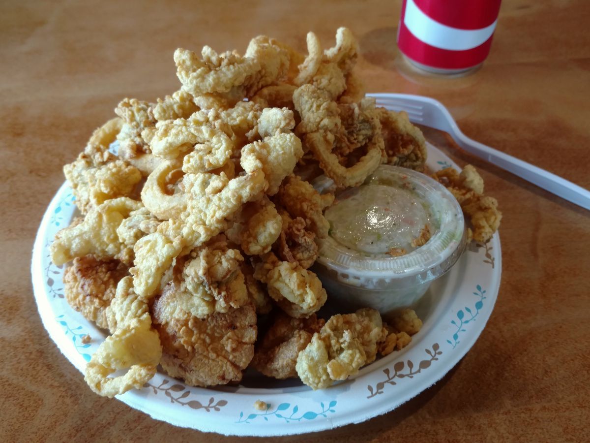 A white paper plate is stacked high with fried seafood with a side of tartar sauce in a plastic cup.