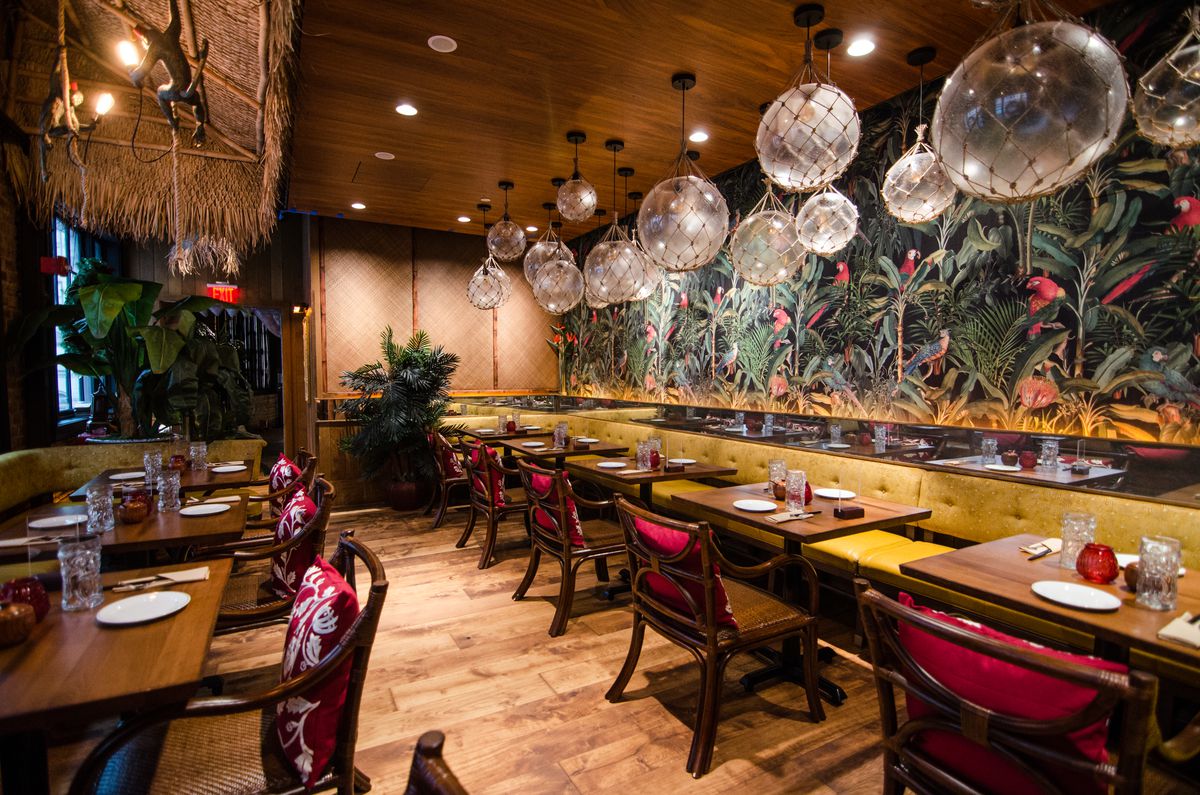 View of the dining room at a tiki-themed restaurant, including tropical wallpaper and big glass dome lighting, as well as a thatch overhang with brass monkey lamps dangling.