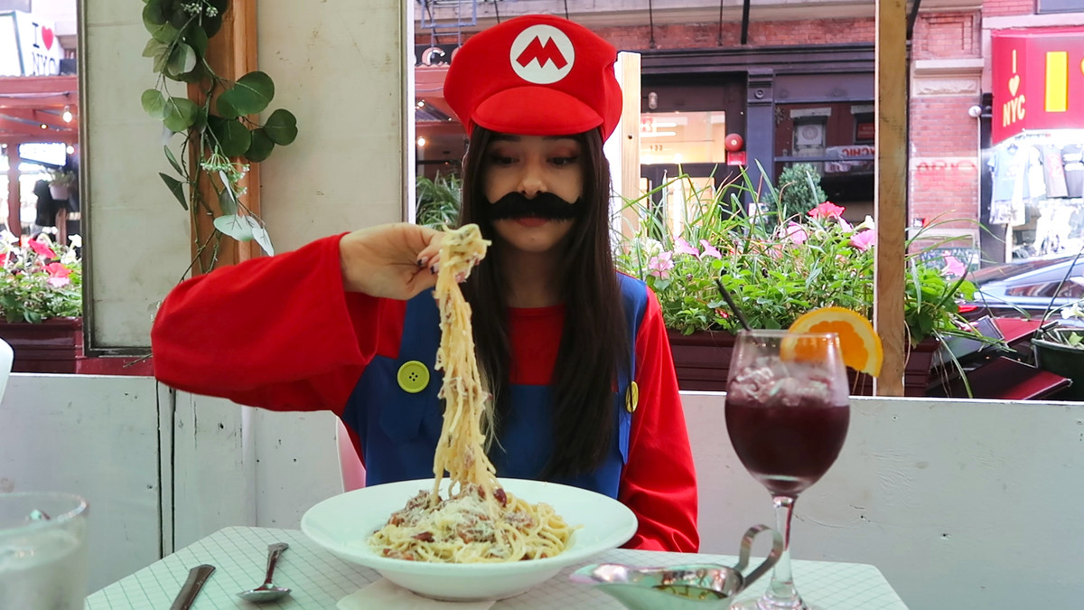 Tina holds a massive forkful of spaghetti, with noodles falling off the fork. She’s dressed as Mario, sitting in an outdoor dining shed on a sunny, flower-lined New York street.