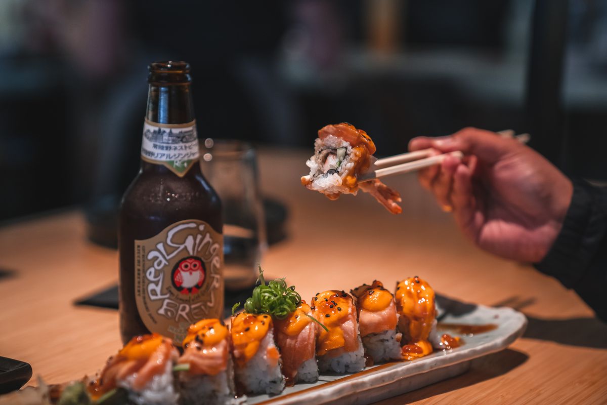 A sushi roll, glass brown beer bottle, a hand holding chopsticks holding a piece of sushi roll from TigerLily in Ferndale, Michigan.