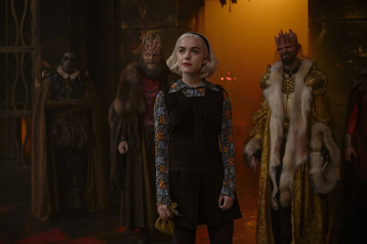  Cute, blonde Sabrina stands in a red-lit room in front of two demons dressed in heavy furs and sporting elaborately bony heads that look like crowns.