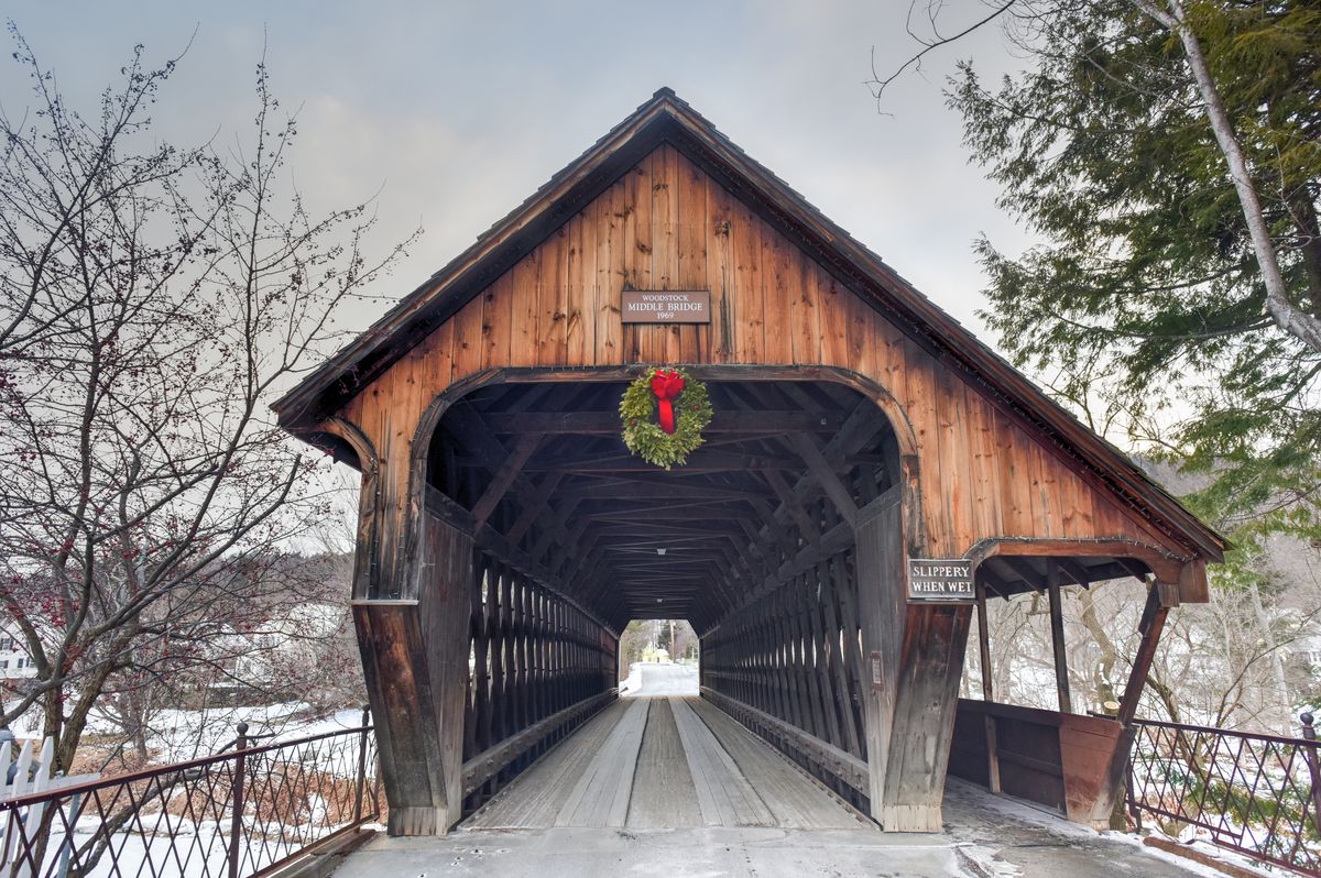 A covered bridge with snowfall in the background and a red and green holiday wreath hanging from the top of the bridge.