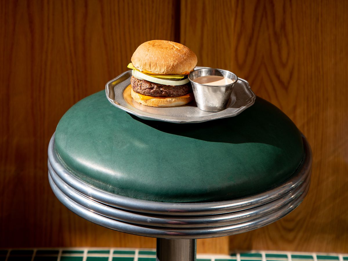 On a green diner stool, a burger on a plate sits with a side of sauce.