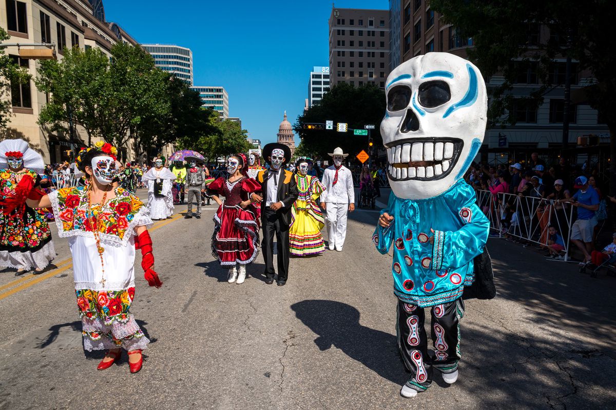 A city street with people dressed in traditional clothing for Dias de los Muertos, including people with their faces painted as skeletons and wearing wedding dresses and suits. One person has on an oversized skeleton puppet head.