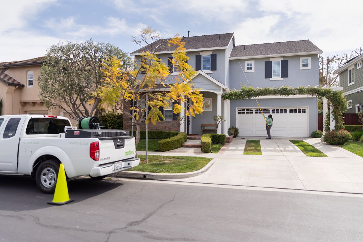 A pest control specialist wearing a green shirt and gray pants uses a yellow pest control wand on the exterior of a white garage door on a gray house. Seen on the street is a white Aptive pick-up truck with other pest control tools in the bed.