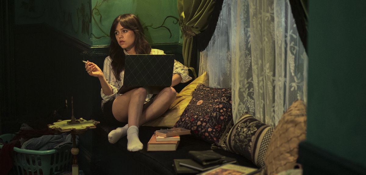 Cairo (Jenna Ortega) sits and smokes with a laptop on her lap and her legs folded up into a chair, surrounded by pillows, books, and a gauzy lace curtain in Miller’s Girl