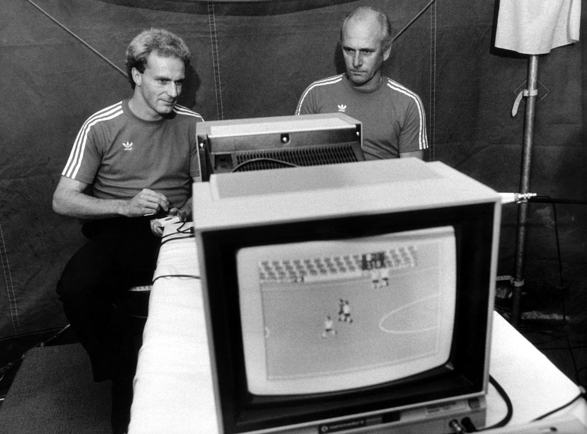 Udo Lattek y Karl-Heinz Rummenigge sentados frente a una computadora en 1983, viendo un viejo videojuego de fútbol en la pantalla.