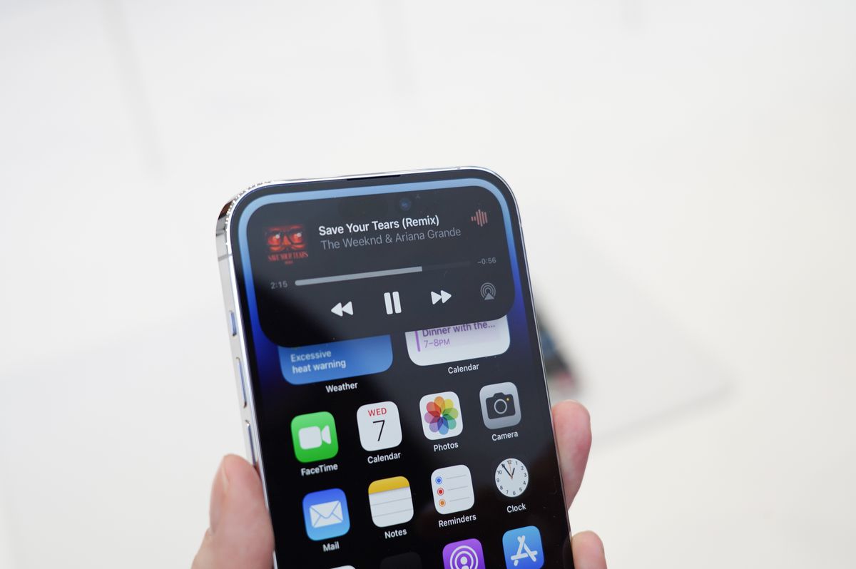 An image of an iPhone held in a hand. The Dynamic Island feature is prominent on the display. Its a black box which shows a music player in it.