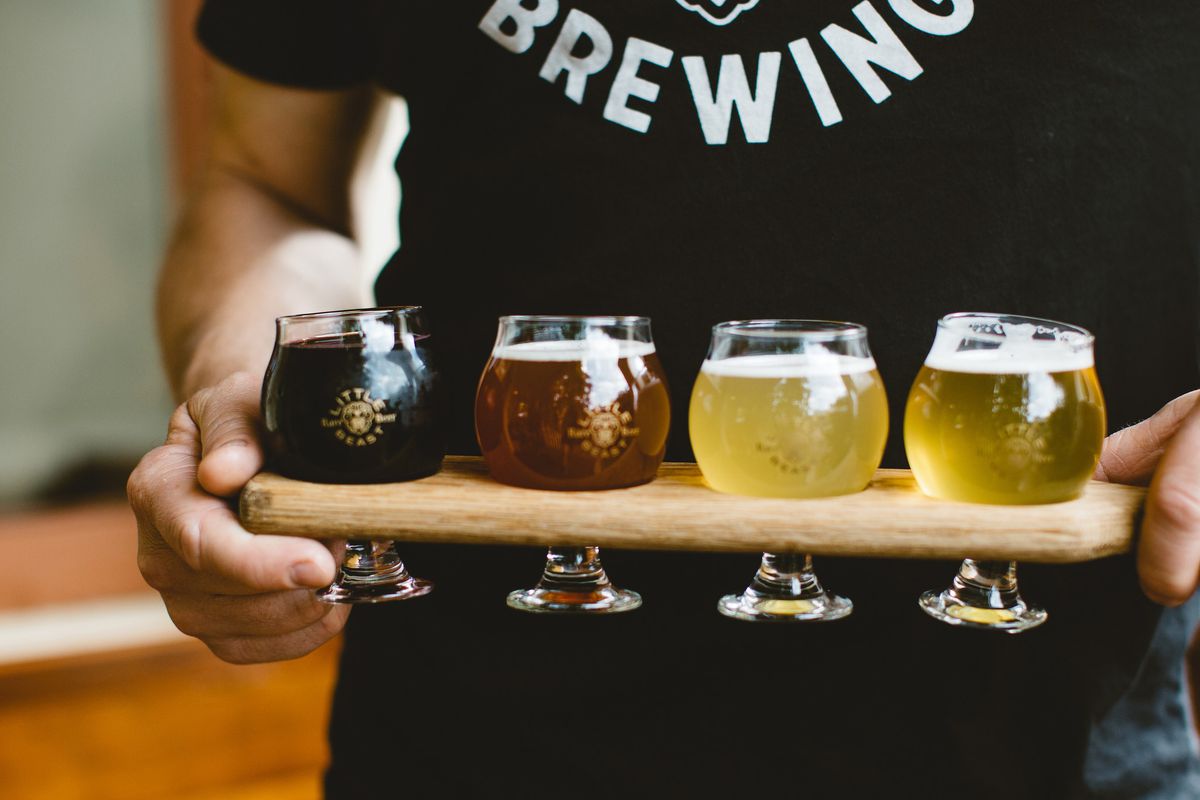 A man in a Little Beast shirt holds a tasting tray with four small glasses of beer, ranging from pale yellow to black.
