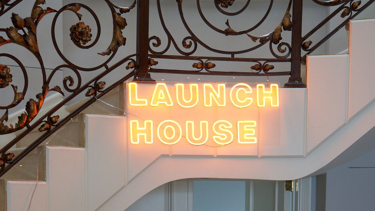 A neon sign that reads “Launch House” mounted on the side of an indoor staircase.