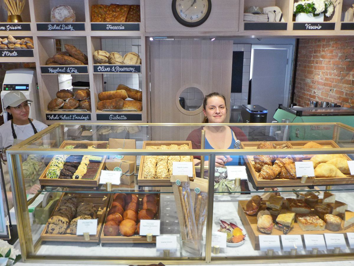 A woman stands behind a pastry counter as two clerks look on.