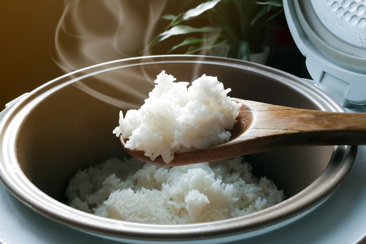 An open rice cooker filled with steaming rice and a wooden spoonful of rice hovers above it