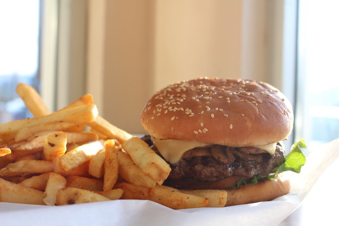 A mushroom swiss burger sits on a plate with fries on a sunny day with a window in the background. The background is too blurry to make out what is outside.