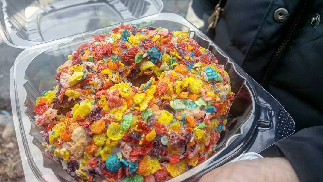 Closeup on a giant doughnut covered with Fruity Pebbles in a plastic takeout container.