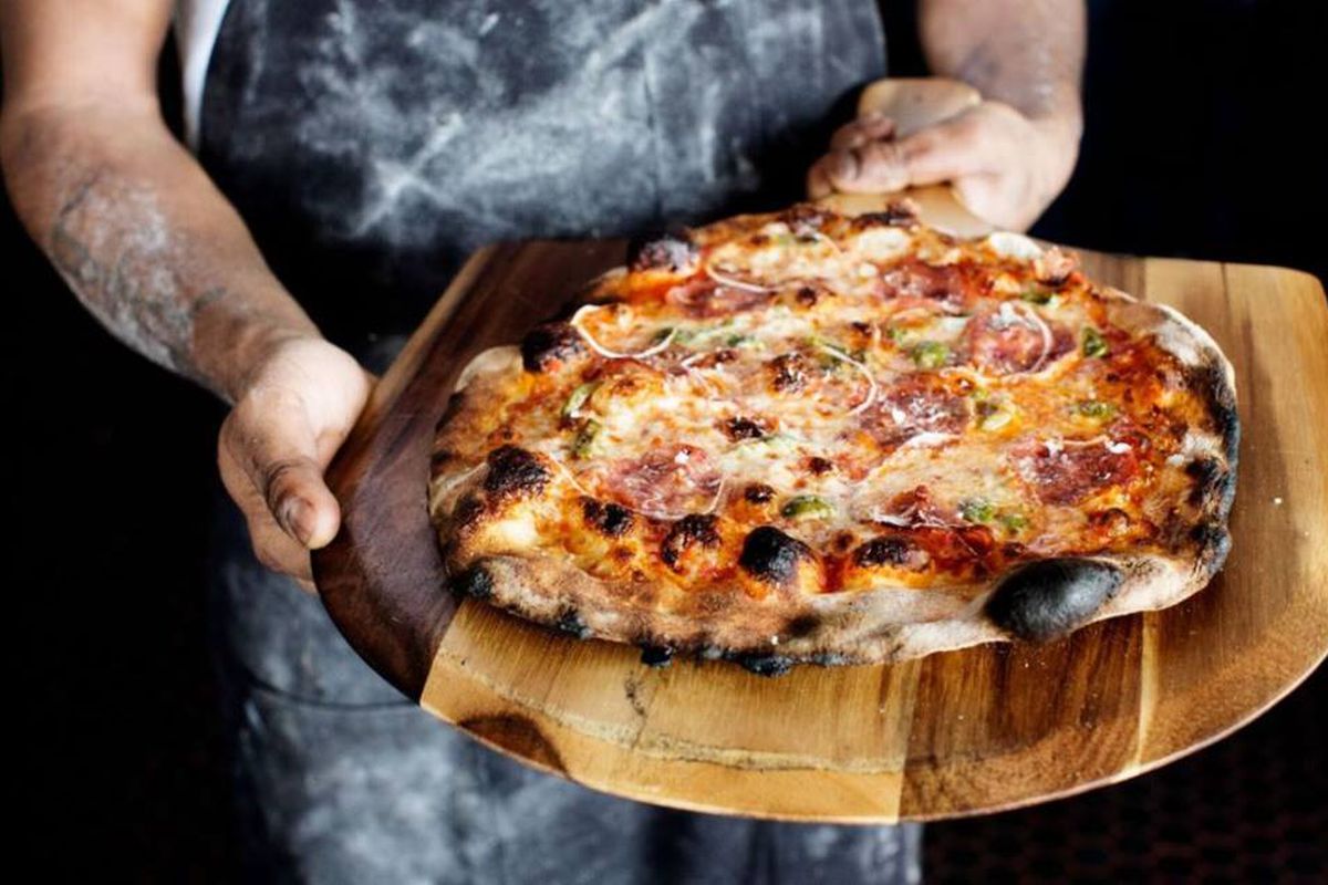 A flour-covered chef presents a blistered crust with cheese and toppings