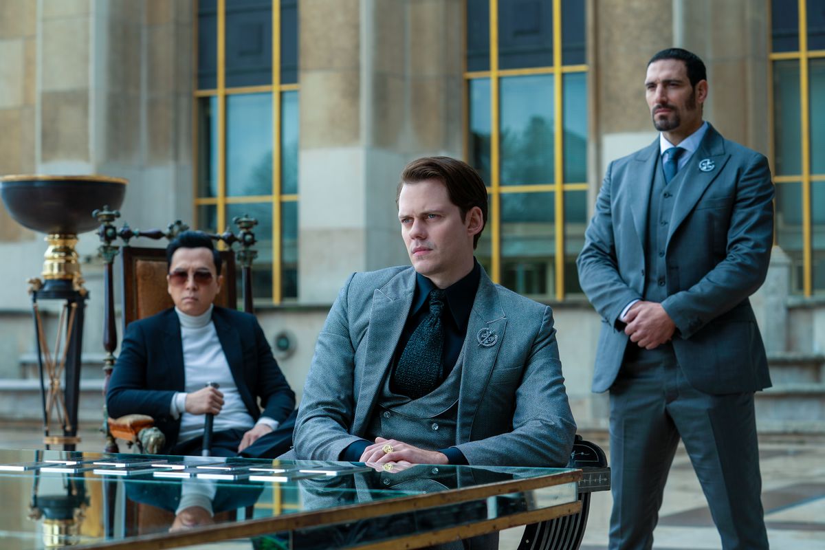 Donnie Yen as Caine sitting in a chair behind Bill Skarsgård as Marquis, who sits at a glass table, guarded by Marko Zaror as Chidi in John Wick: Chapter 4