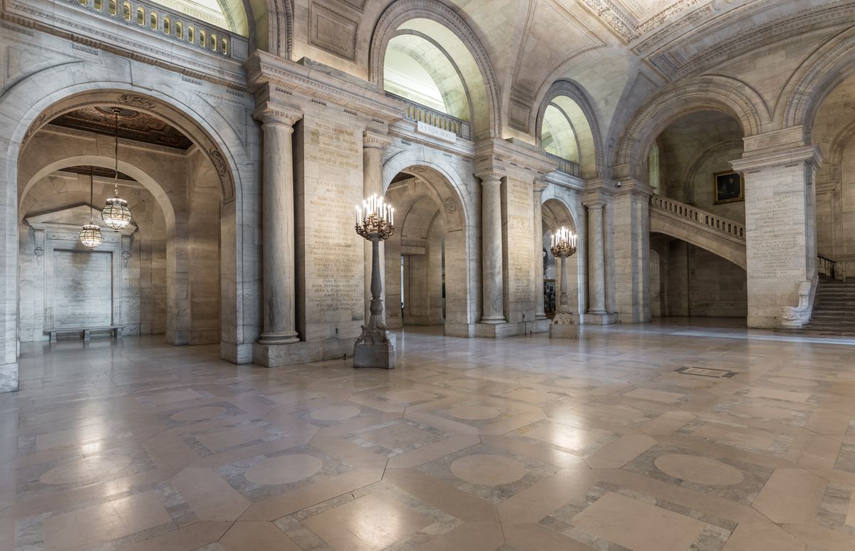 The interior of the New York Public Library. There are arches and candelabras along the walls and a large staircase.