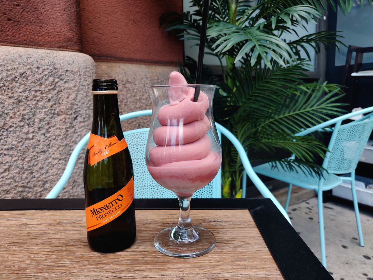 A glass sits on a restaurant patio table filled with a swirl of pale pink soft serve, with a split of sparkling wine to the side