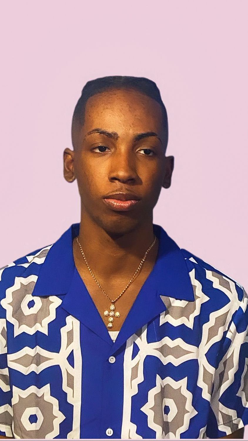 Zadane Russell stands for a portrait against a light purple background, wearing a blue, white and grey patterned shirt and a cross necklace.