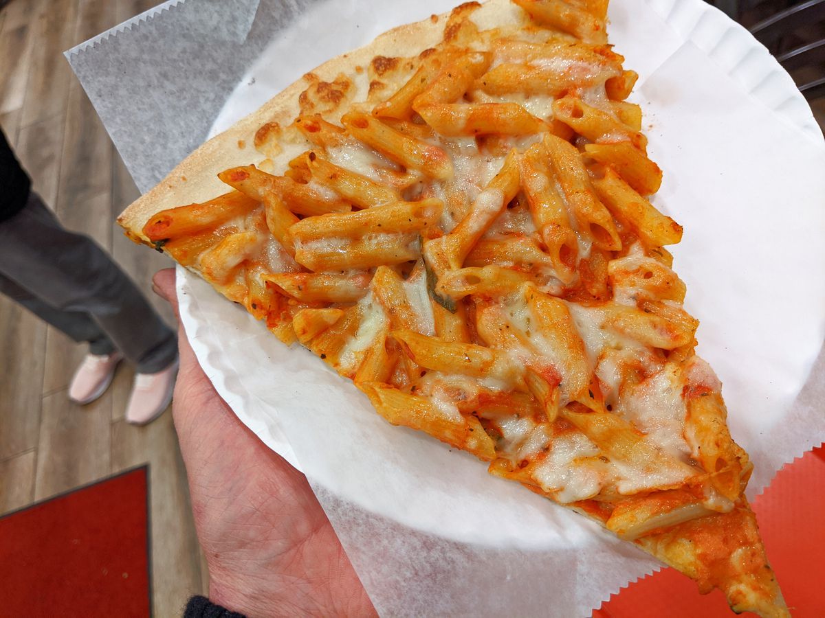 A ziti slice of pizza held aloft on a white paper plate