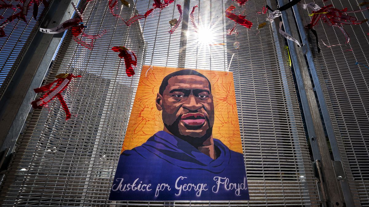 A painting of George Floyd reading “justice for George Floyd” hangs on a fence made of tight mesh. Red ribbons have been tied around the painting; these flap in the breeze, as the sun shines above the entire scene.