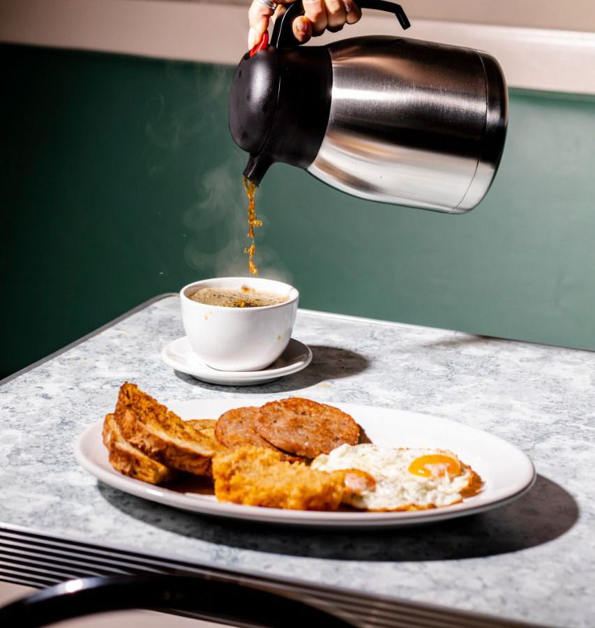 An unseen hand pours coffee into a mug beside a plate of mixed breakfast items. 