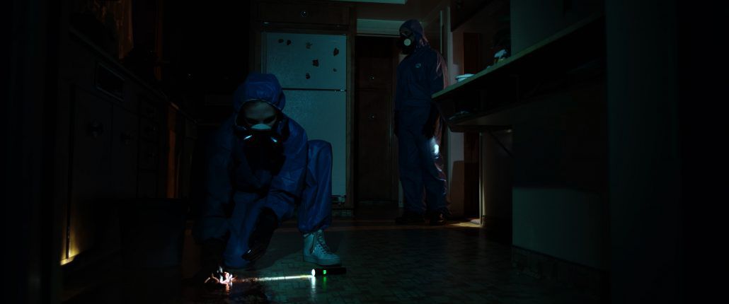 Two figures in hazmat suits inspect the floor of a darkened kitchen in 11th Hour Cleaning.