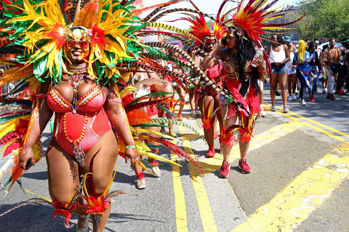 Women in colorful costumes with feathered headdresses walk down a street during a large neighborhood parade. 