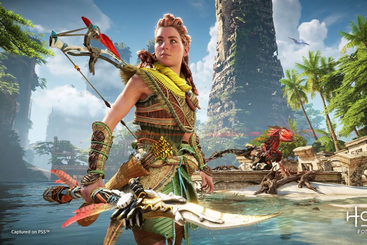 Aloy standing on a beach holding a spear looking into the distance while a Machine prowls in the background in Horizon Forbidden West