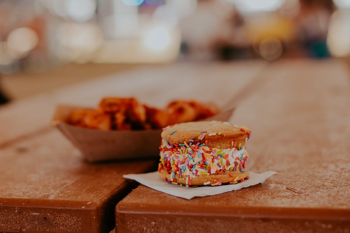 An ice cream sandwich made of cookies and rainbow sprinkle-covered vanilla ice cream on a picnic table.