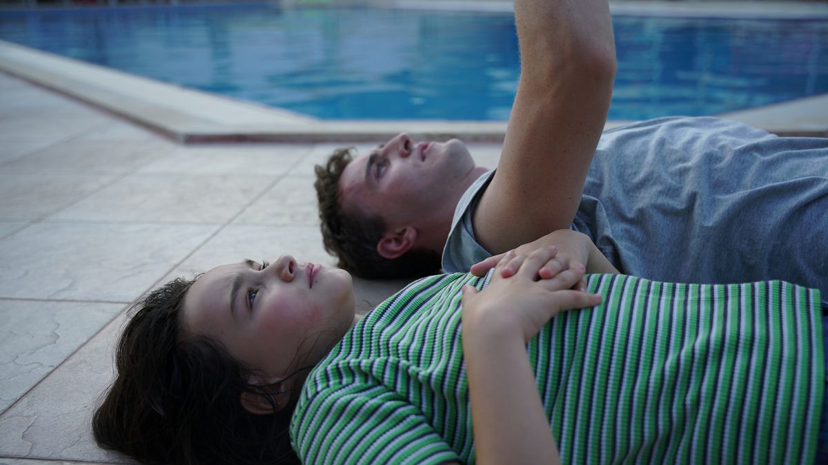 A father and daughter lie on their backs next to a pool while the father points at the sky in Aftersun.