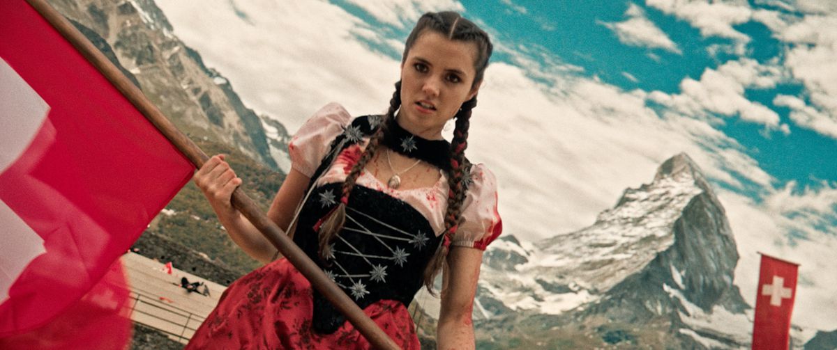Alice Lucy as Heidi, dressed in a blood-splattered dress, holding a red flag with the Swiss Alps in the background in Mad Heidi.
