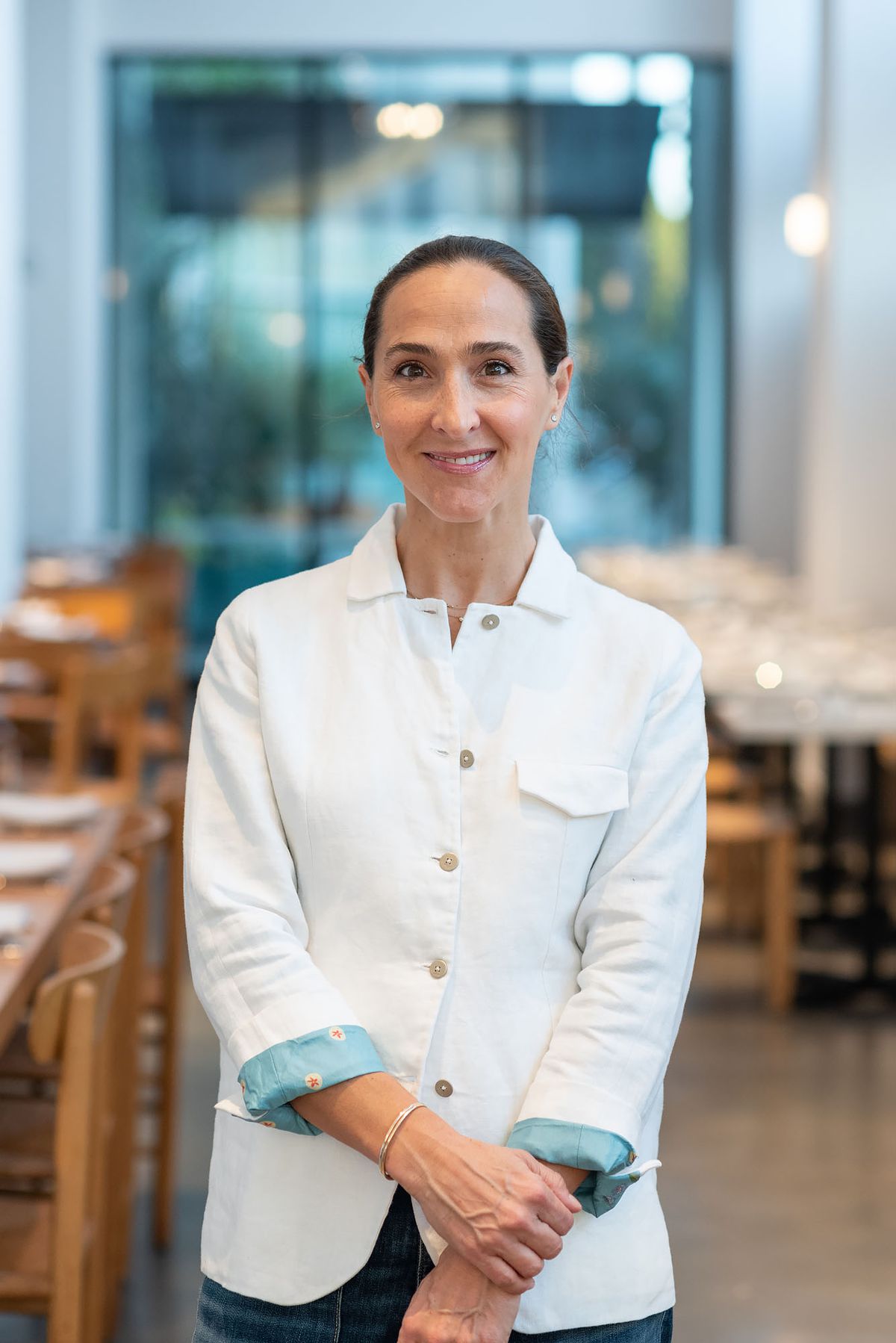 A woman in a chef’s coat, her hair pulled back, holds one wrist and smiles.