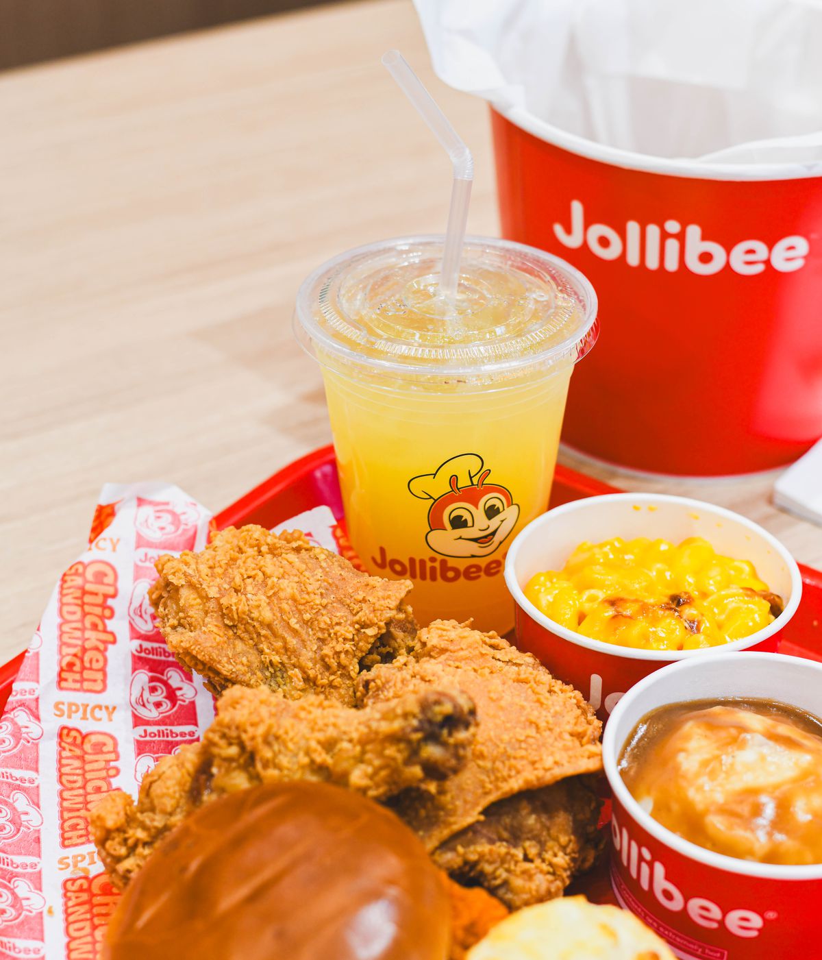 Fried chicken, mashed potatoes and gravy and mac and cheese in red and white cups, a yellow liquid in a clear cup with a red bee wearing a chef hat and Jollibee in red printed on it and a bun set on a red tray with multi-colored liner and a red and white Jollibee bucket set next to it on a light surface.