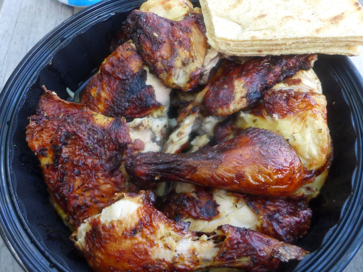 A black plastic container filled with well browned chicken parts, with cut pitas on the side.