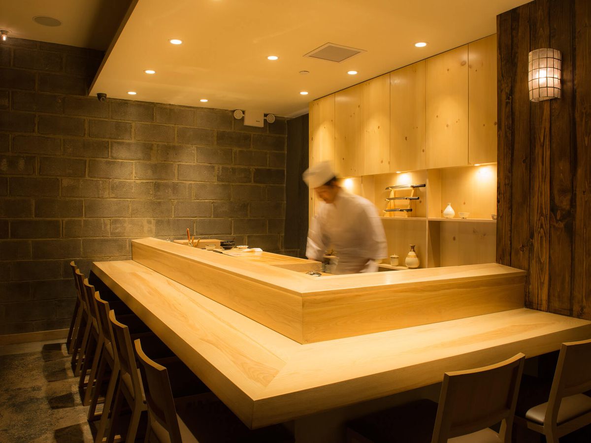 Sushi Amane’s sushi bar counter has yellow wood, with a chef wearing white standing in the middle.