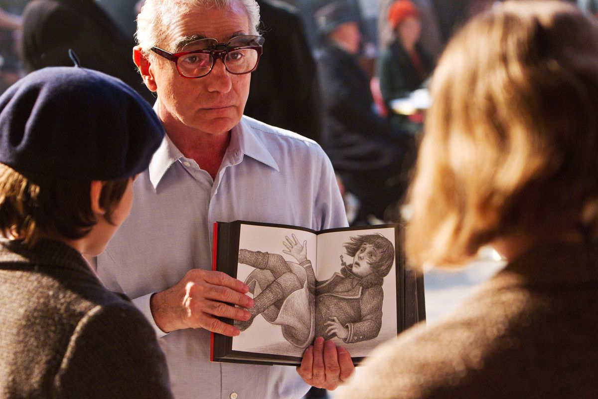 Martin Scorsese holds up an open book to show a black-and-white drawn image of a surprised-looking child with shoulder-length hair falling down to actor Asa Butterfield and a second, unidentified figure, both with their backs to the camera, on the set of 2011’s Hugo