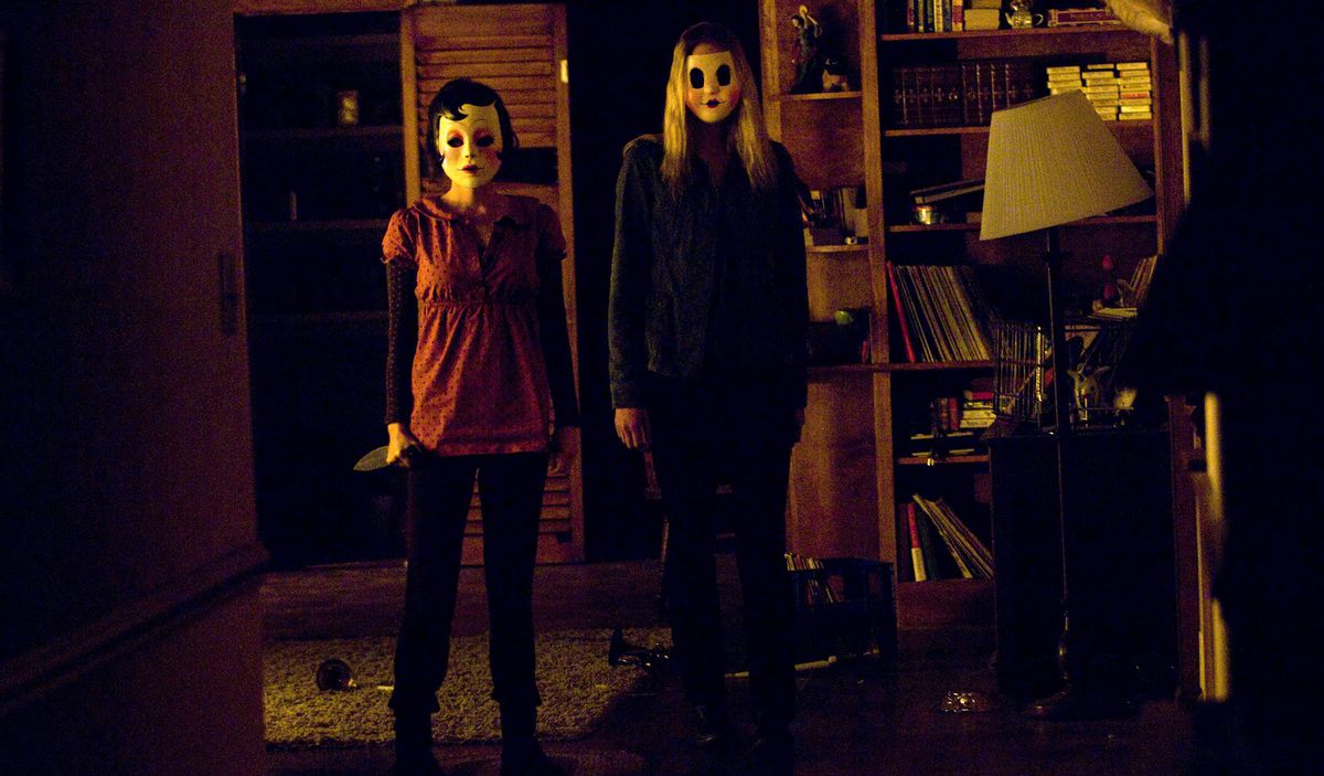 Two figures in stylized girl masks with blush on the cheeks and faint smiles loom alarmingly in a cluttered cabin room in The Strangers