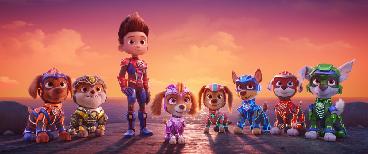 A boy in an armored suit flanked by several dogs in similar armored suits standing on a cliffside overlooking an ocean and sunset sky in Paw Patrol: The Mighty Movie.