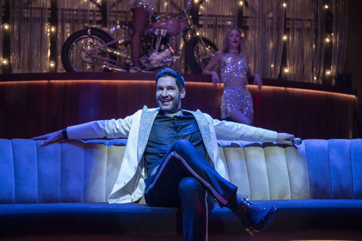 Tom Ellis as Lucifer sits smugly on a couch under blue light in season 6 of Lucifer