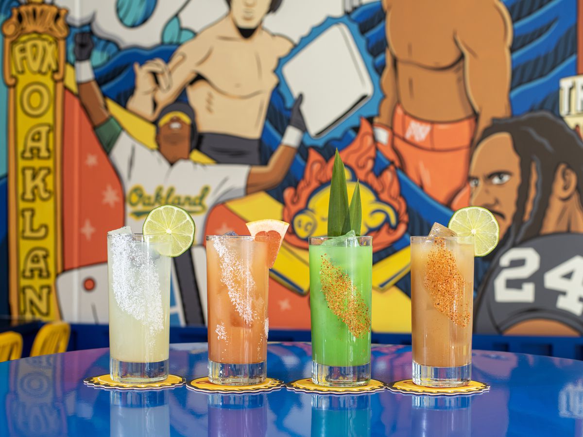 Four glasses with drinks of different colors, in front of a mural depicting heroes of Oakland at Senor Sisig.