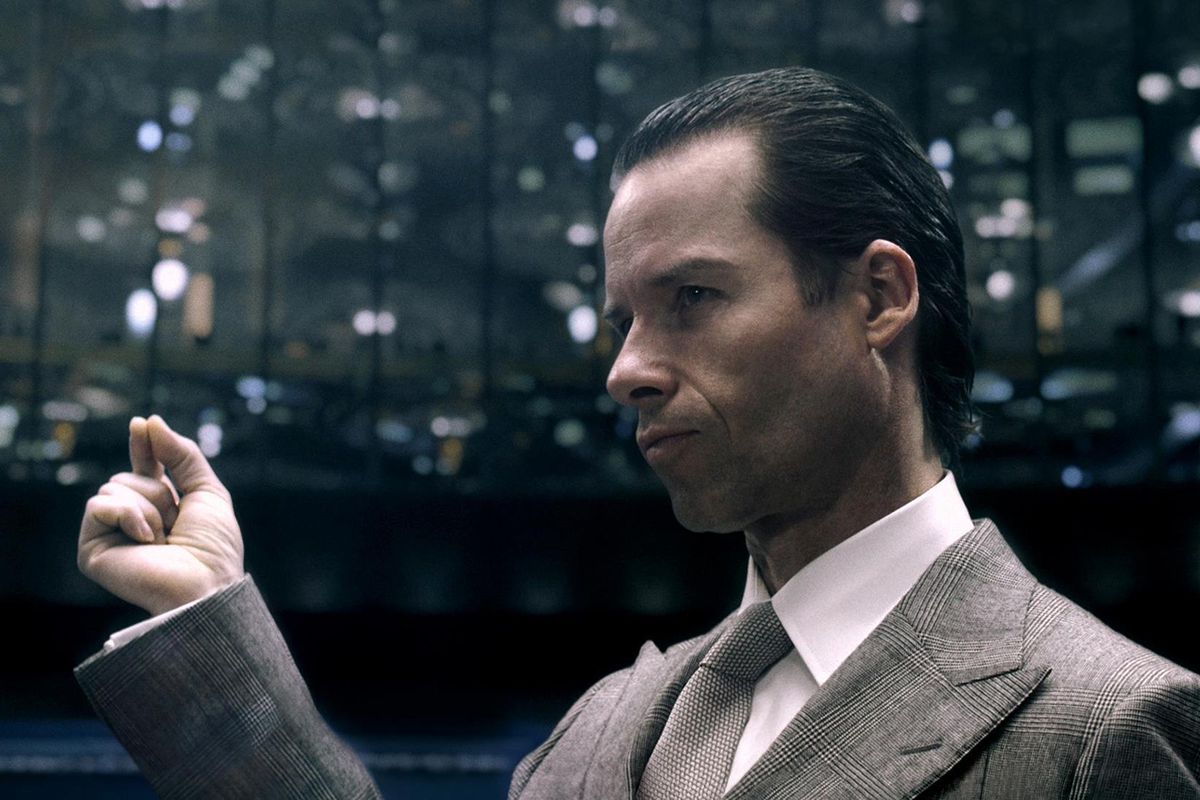 A close-up shot of Peter Weyland (Guy Pearce) in a tan suit and tie pinching his forefinger and thumb together while surrounded by an auditorium of people in a fake TED talk produced for the 2012 film Prometheus.