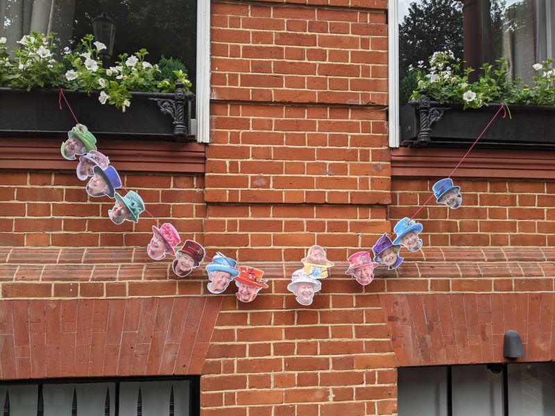 A decorative garland of photos of the queen wearing various multicolored hats, hanging outside a brick building.